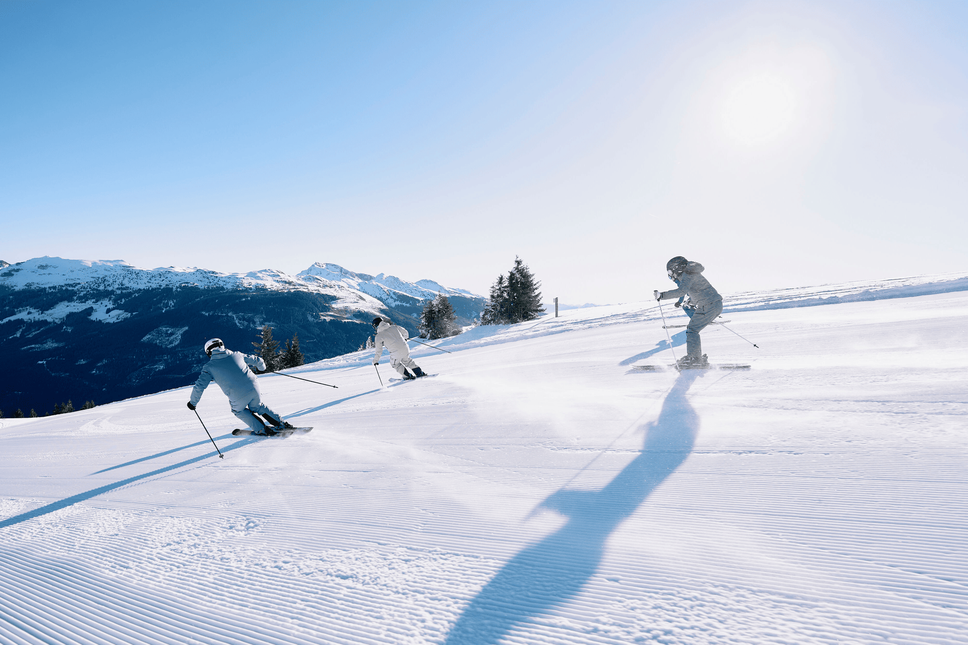 Three skiers descend a sunlit slope, casting long shadows on the pristine snow. Snowy mountains and trees frame the scene under a clear blue sky.