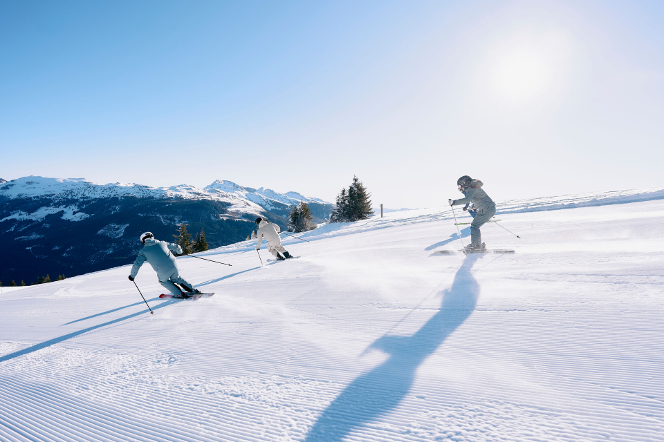 Three skiers descend a sunlit slope, casting long shadows on the pristine snow. Snowy mountains and trees frame the scene under a clear blue sky.