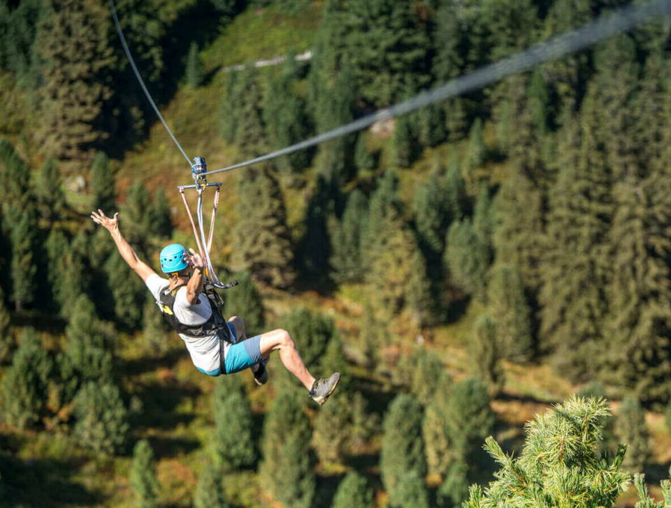 Eine Person mit Helm und Klettergurt gleitet mit ausgestreckten Armen voller Begeisterung über einen dichten Wald hinweg. Sonnenlicht fällt durch die hohen immergrünen Bäume.