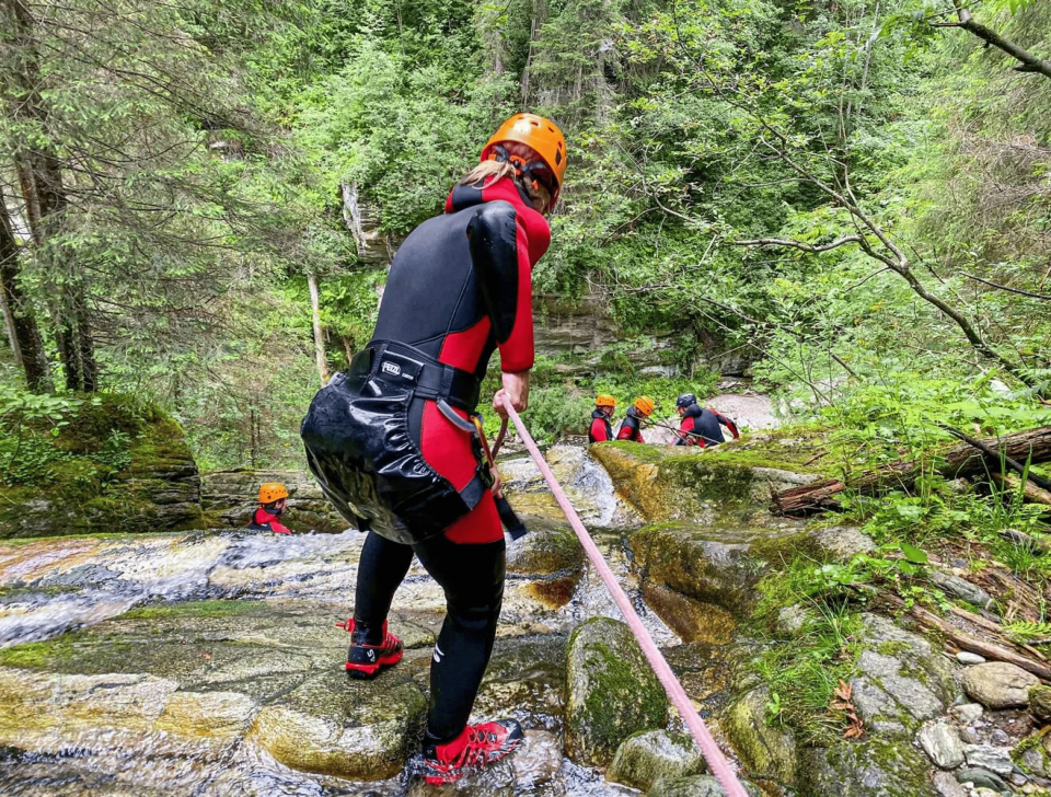 Eine Person in Neoprenanzug und Helm seilt sich in einem üppigen Wald an einem Wasserfall ab. Andere in ähnlicher Ausrüstung schauen zu und vermitteln Abenteuerlust und Teamgeist.