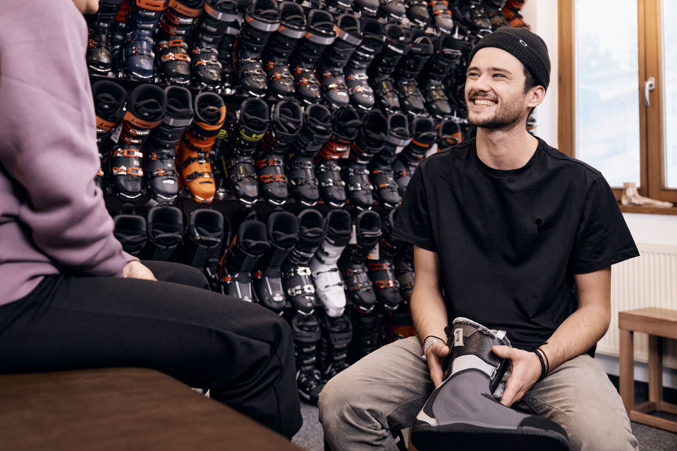 Ein lächelnder Mann hält einen Skischuh in einem Geschäft in der Hand, umgeben von einer Wand voller Skischuhe. Er trägt Freizeitkleidung und eine Mütze. Die Atmosphäre ist freundlich und einladend.
