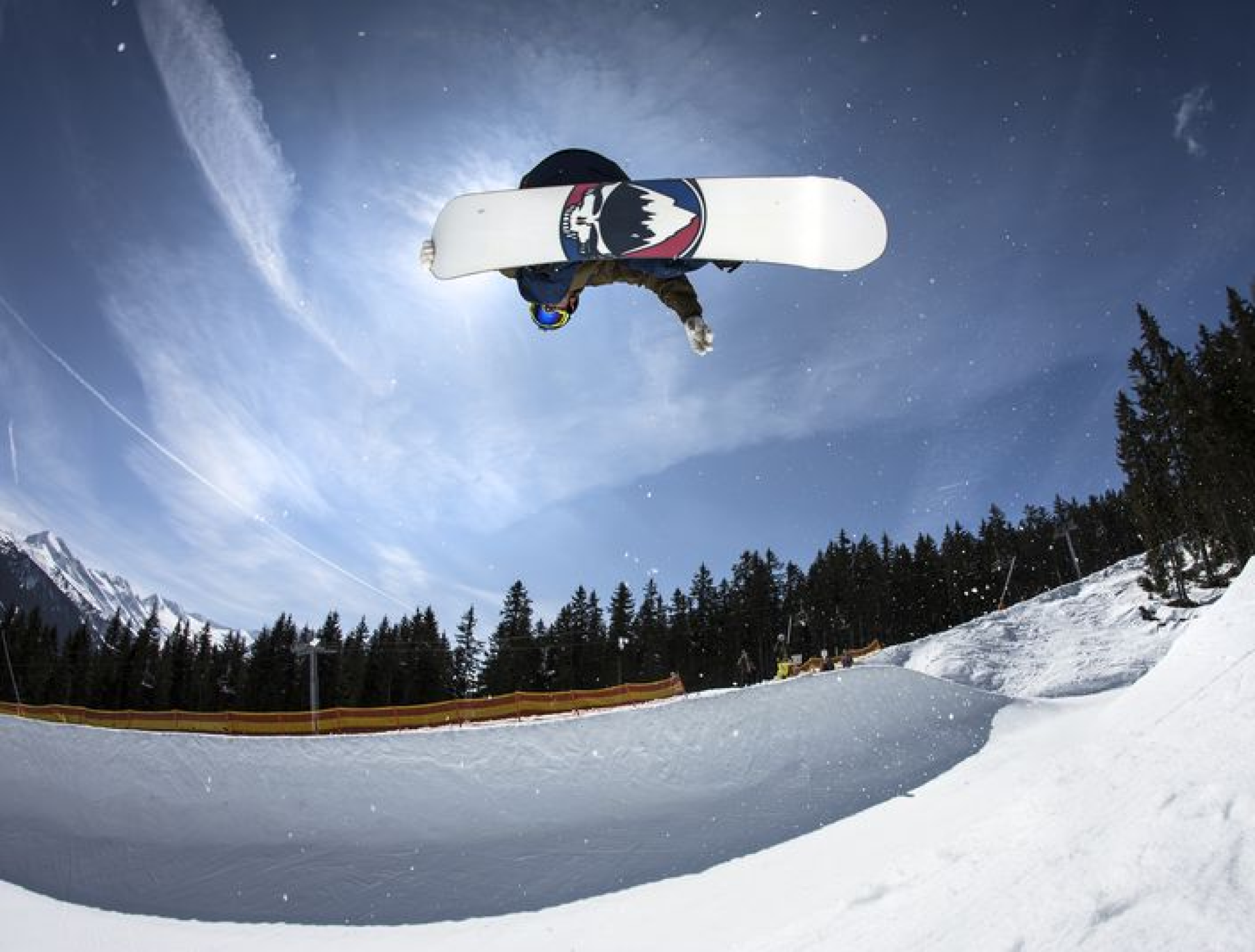 Snowboarder, der vor strahlend blauem Himmel einen Sprungtrick ausführt. Umgeben von verschneiten Hängen und Kiefern, fängt er Action und Abenteuer ein.