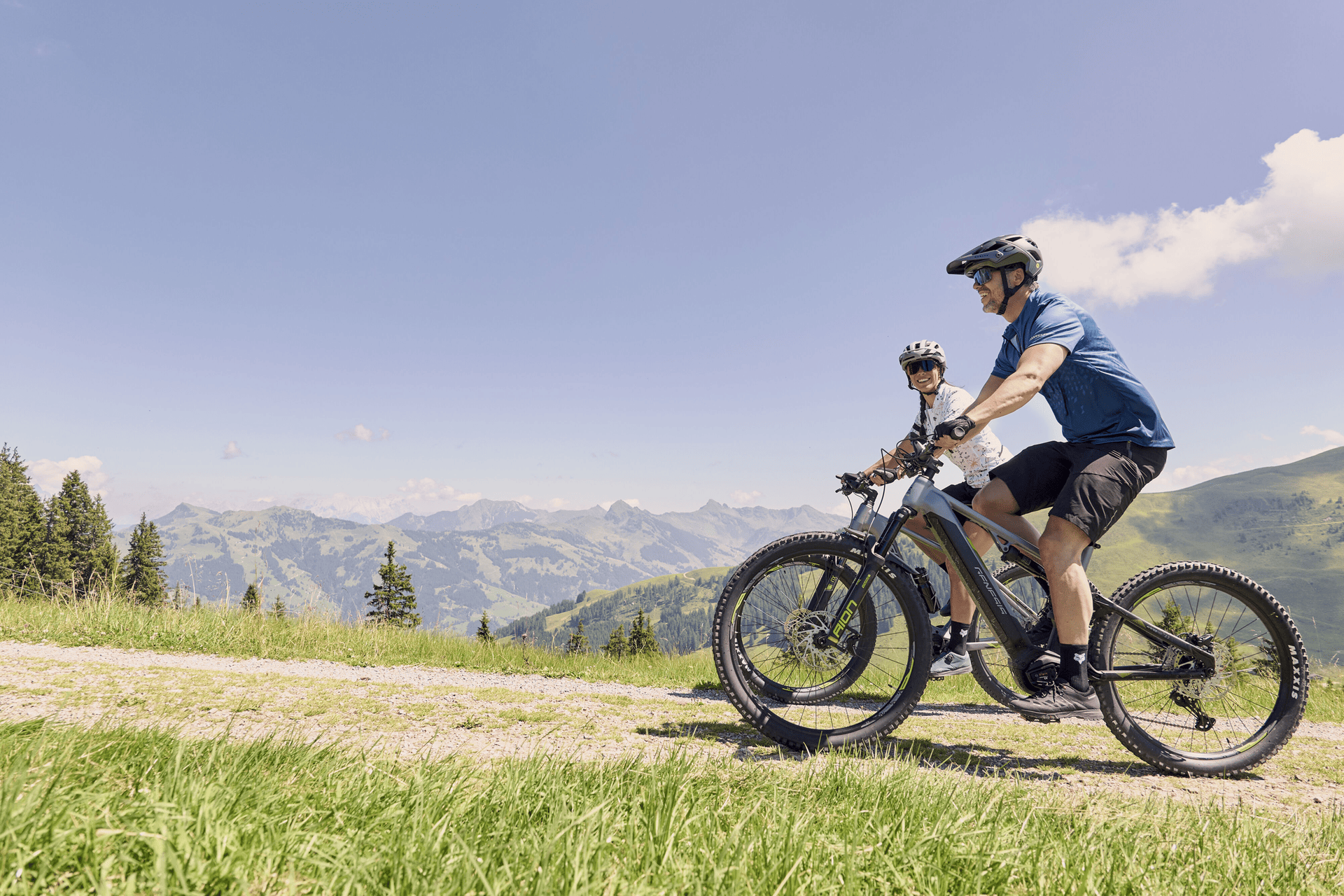Zwei Radfahrer fahren mit Mountainbikes auf einem malerischen Weg, umgeben von üppig grünen Hügeln und fernen Bergen, unter einem strahlend blauen Himmel.
