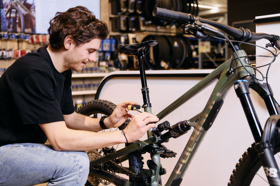 Ein Mann in schwarzem Hemd und Jeans justiert in einem Geschäft ein grünes Mountainbike und konzentriert sich dabei auf die Gangschaltung. Der Schauplatz ist ein gut beleuchteter, übersichtlicher Fahrradladen.