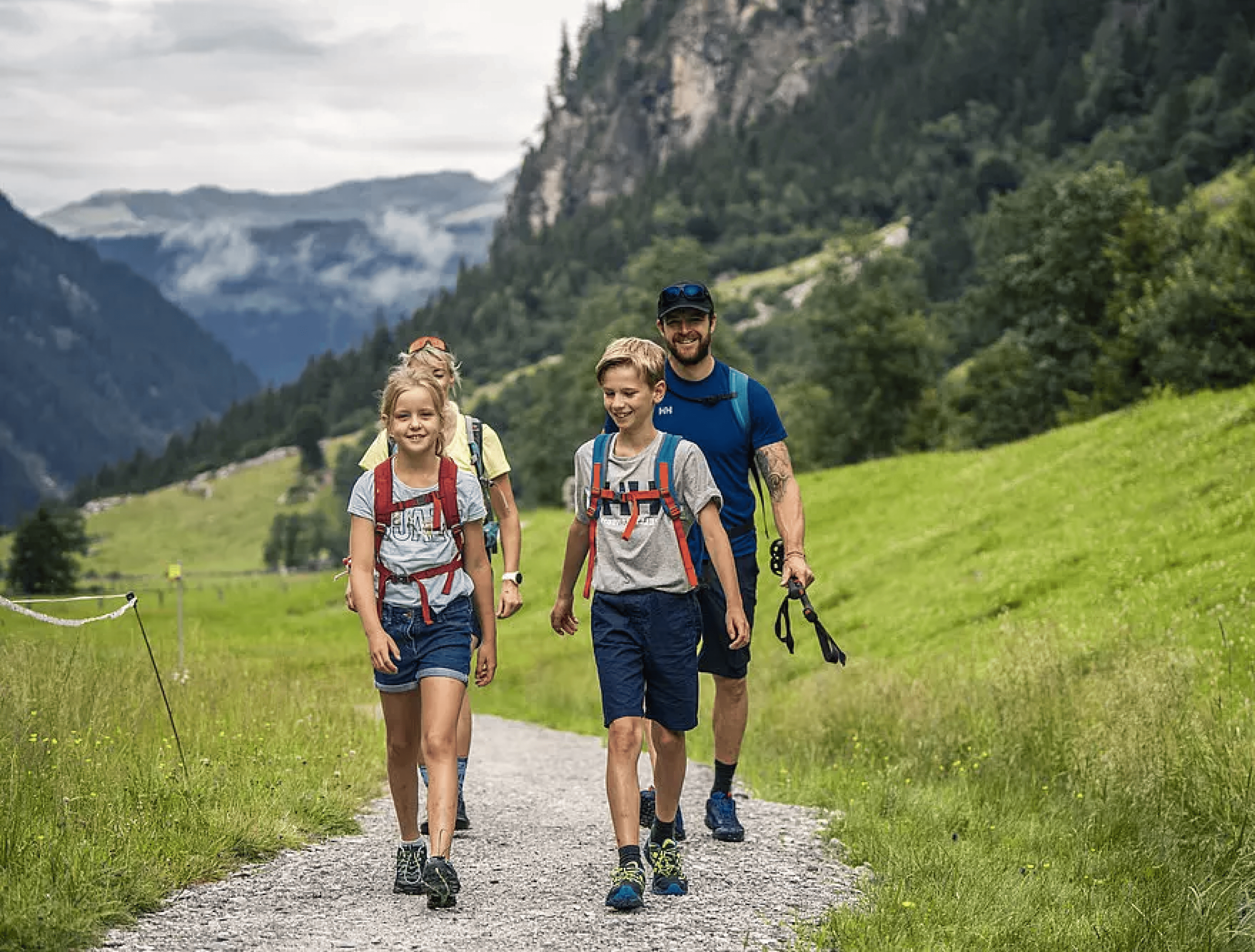 Eine glückliche vierköpfige Familie wandert auf einem Bergpfad, umgeben von üppigem Grün und hoch aufragenden Gipfeln. Der Himmel ist bewölkt und schafft eine ruhige Atmosphäre.