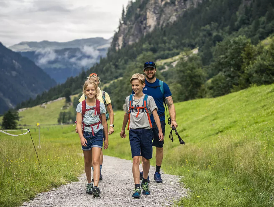 Eine glückliche vierköpfige Familie wandert auf einem Bergpfad, umgeben von üppigem Grün und hoch aufragenden Gipfeln. Der Himmel ist bewölkt und schafft eine ruhige Atmosphäre.