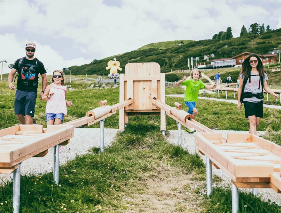 Eine vierköpfige Familie spielt begeistert auf einer hölzernen Wasserspielanlage auf einer Wiese mit Hügeln und einigen Gebäuden im Hintergrund unter einem bewölkten Himmel.