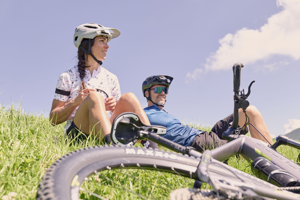 Eine Frau und ein Mann in Fahrradkleidung entspannen sich auf einem grasbewachsenen Hügel unter klarem Himmel, mit Fahrrädern in der Nähe, und vermitteln ein Gefühl von Muße und Abenteuer.