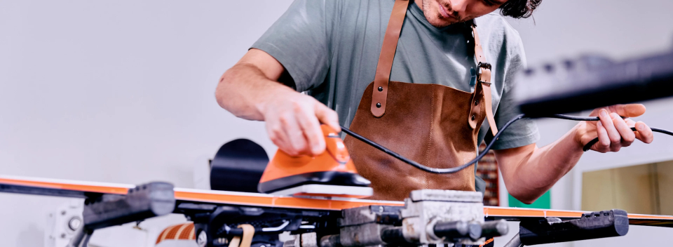 A focused man in an apron waxes a ski in a workshop. He uses an iron, conveying precision and dedication. The atmosphere is professional and serious.