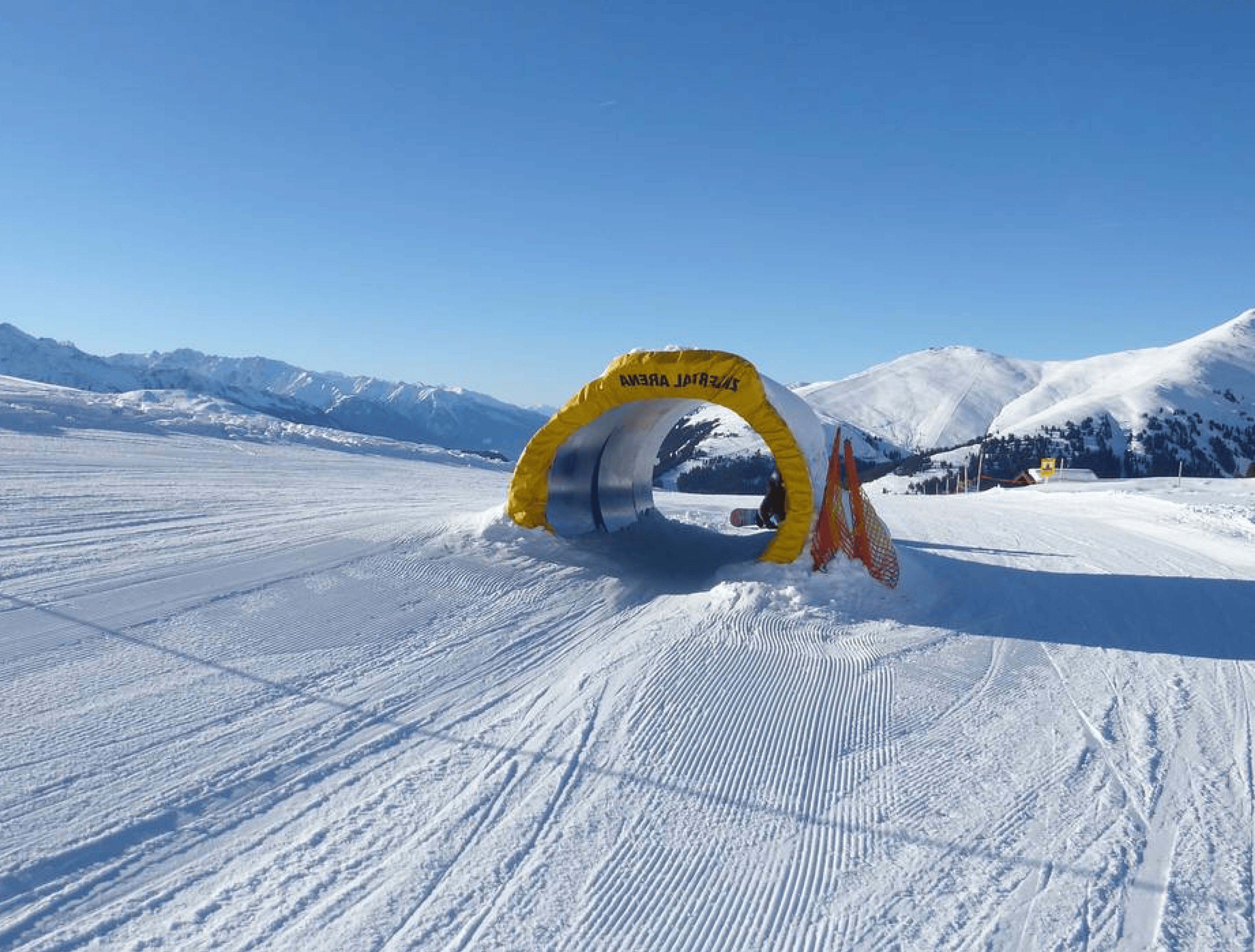 A snowy ski slope with a yellow tunnel labeled "SMILE AGAIN" under a clear blue sky. Mountains in the background create a serene, inviting atmosphere.