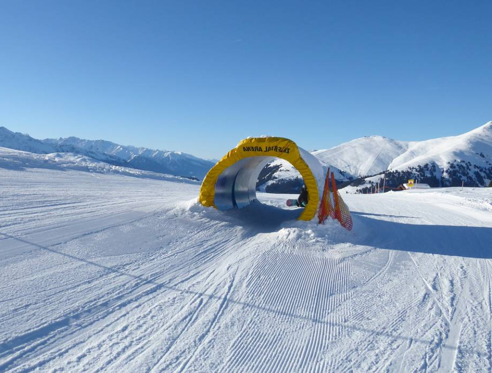 A snowy ski slope with a yellow tunnel labeled "SMILE AGAIN" under a clear blue sky. Mountains in the background create a serene, inviting atmosphere.