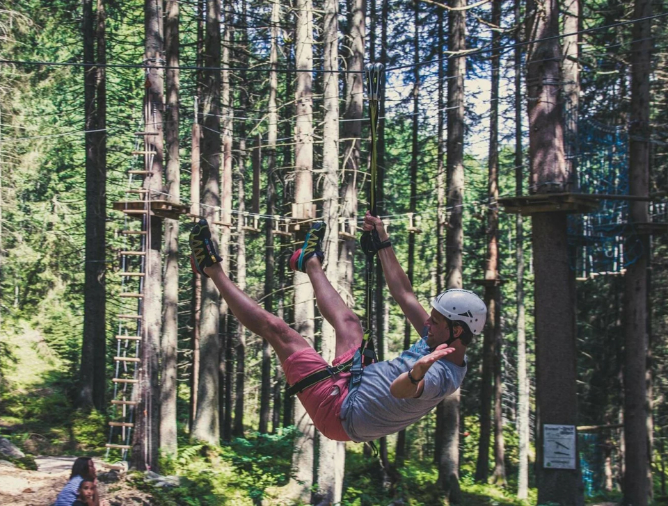 Eine Person saust voller Freude mit einer Seilrutsche durch einen üppigen Wald mit hohen Bäumen. Mit Helm und Klettergurt ausgestattet, vermittelt sie Aufregung und Abenteuer inmitten der Natur.