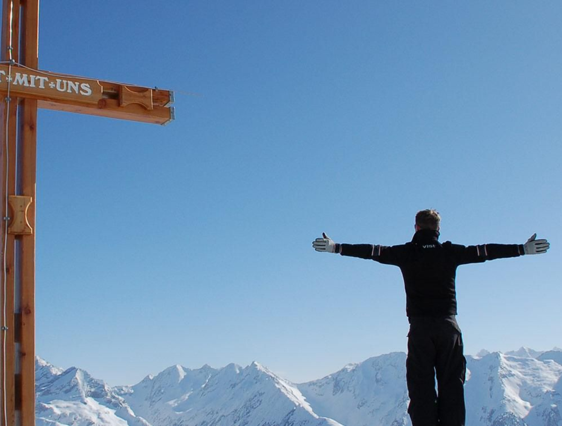 Eine Person in schwarzer Winterkleidung steht mit ausgestreckten Armen auf einem schneebedeckten Berggipfel. In der Nähe befindet sich ein Holzkreuz unter einem klaren blauen Himmel, das Freiheit vermittelt.