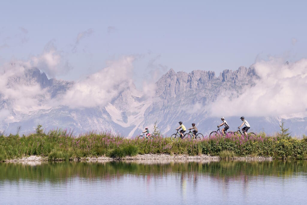 Vier Radfahrer fahren auf einem grasbewachsenen Weg in einer bergigen Landschaft unter einem strahlend blauen Himmel. Sie wirken entspannt und genießen die malerische Aussicht.
