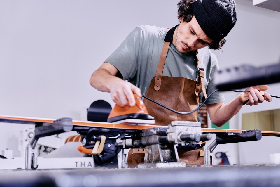 A focused man in an apron waxes a ski in a workshop. He uses an iron, conveying precision and dedication. The atmosphere is professional and serious.