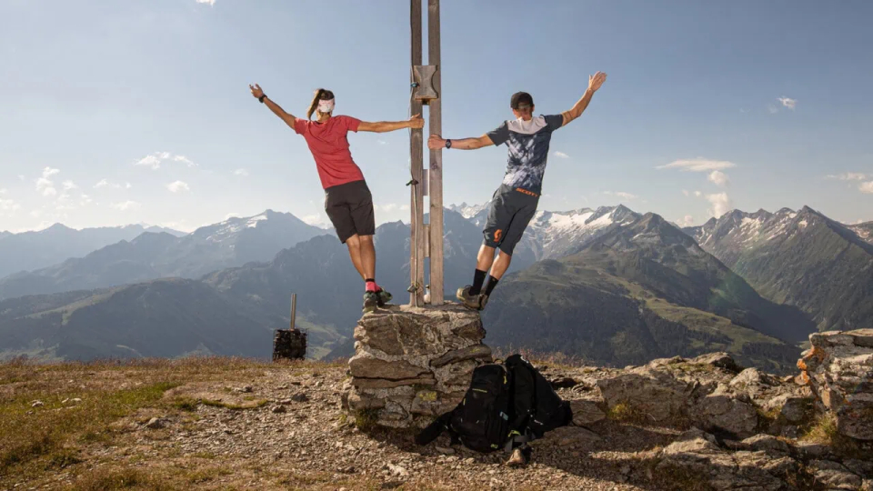 Zwei Menschen balancieren fröhlich auf einem Berggipfel in der Nähe eines Gipfelkreuzes, vor dem Hintergrund ferner blauer Berge und eines klaren Himmels.