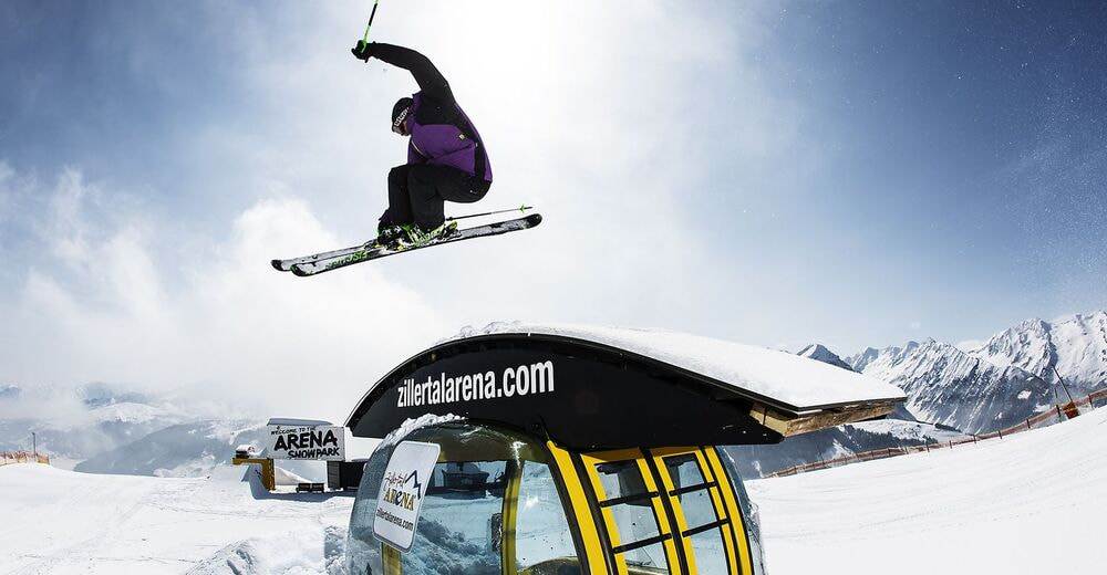 Skier in mid-air doing a trick over a snow-covered gondola with "zillertalarena.com" written on it. Snowy mountains and clear blue sky in the background.