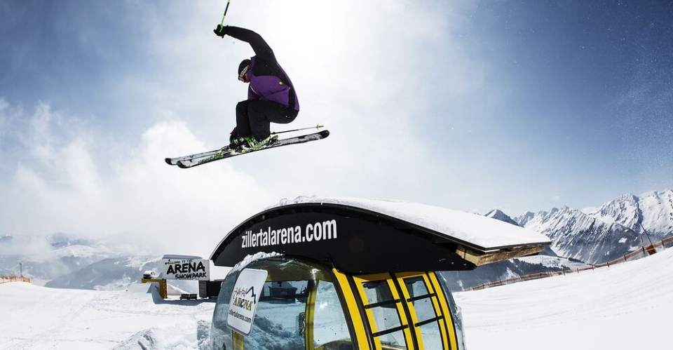 Skier in mid-air doing a trick over a snow-covered gondola with "zillertalarena.com" written on it. Snowy mountains and clear blue sky in the background.