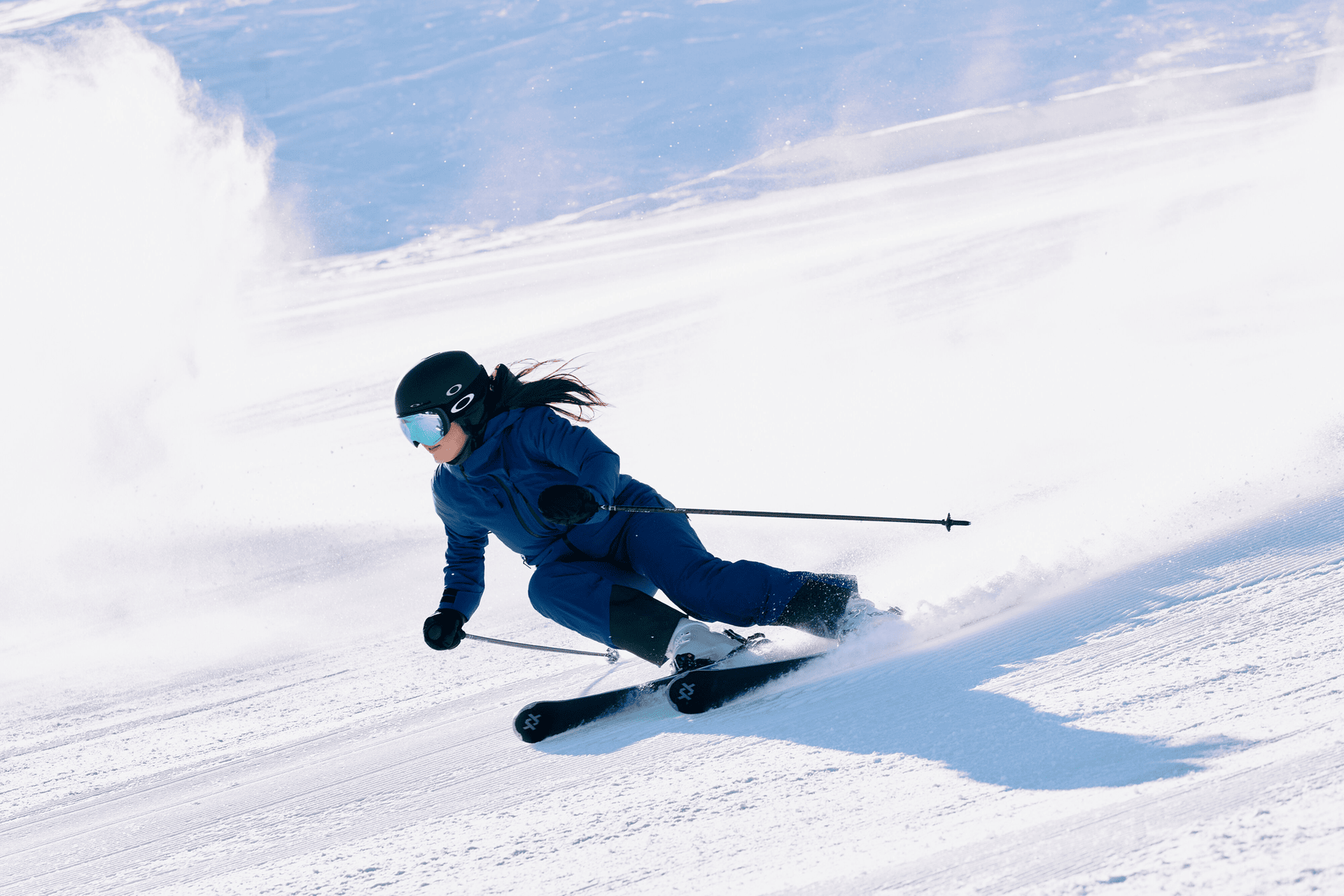 Ein Skifahrer in blauer Kleidung und mit Helm fährt schnell einen verschneiten Hang hinunter und wirbelt dabei Schnee auf. Die Szene vermittelt Geschwindigkeit und Spannung im strahlenden Sonnenlicht.