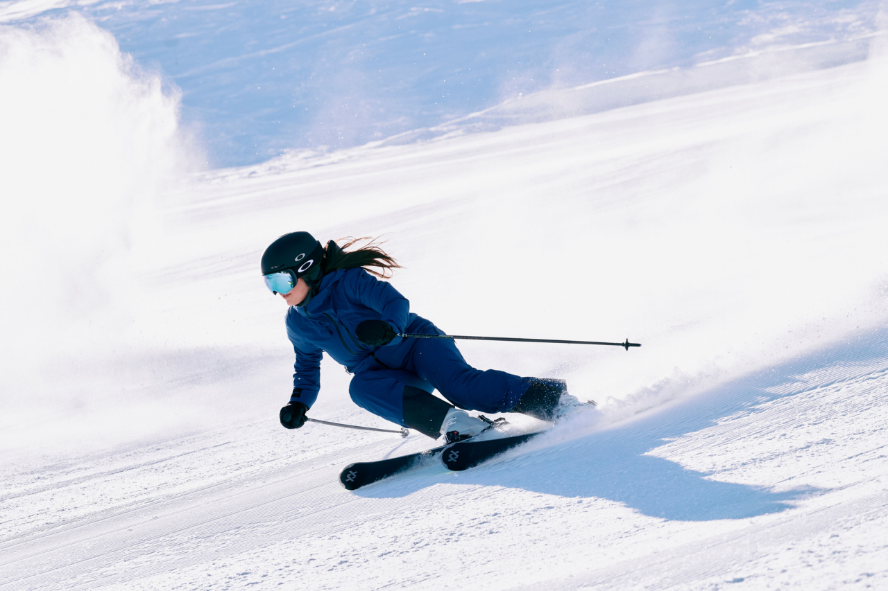 A skier in a blue outfit and helmet is swiftly descending a snowy slope, creating a spray of snow. The scene conveys speed and excitement in bright sunlight.