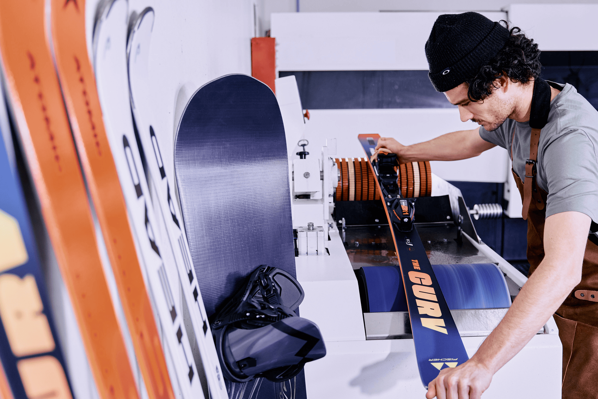 A man works in a ski workshop, using a machine to tune skis. Nearby, various skis and a snowboard are upright, suggesting precision and focus.