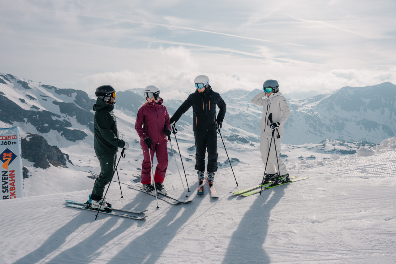Vier Skifahrer in bunten Outfits stehen auf einem schneebedeckten Berggipfel unter einem bewölkten Himmel und unterhalten sich. Majestätische Berge bilden die Kulisse.