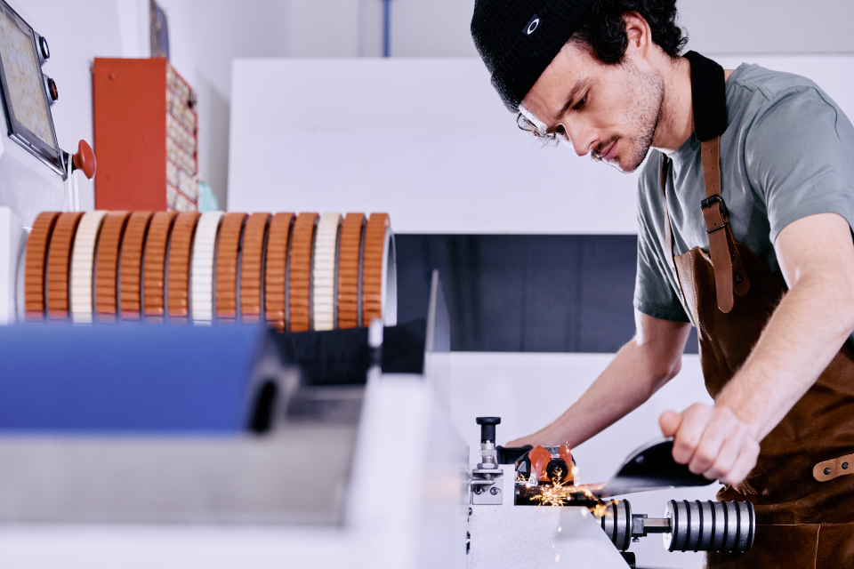 A focused worker wearing an apron operates machinery, adjusting a metal part with precision. The scene conveys concentration and industrial craftsmanship.