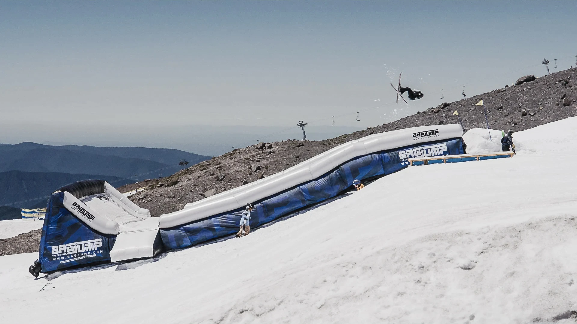 A snowboarder performs an aerial trick above a snowy slope with a blue and white inflatable ramp. Clear sky and distant mountains enhance the scene's adventurous feel.