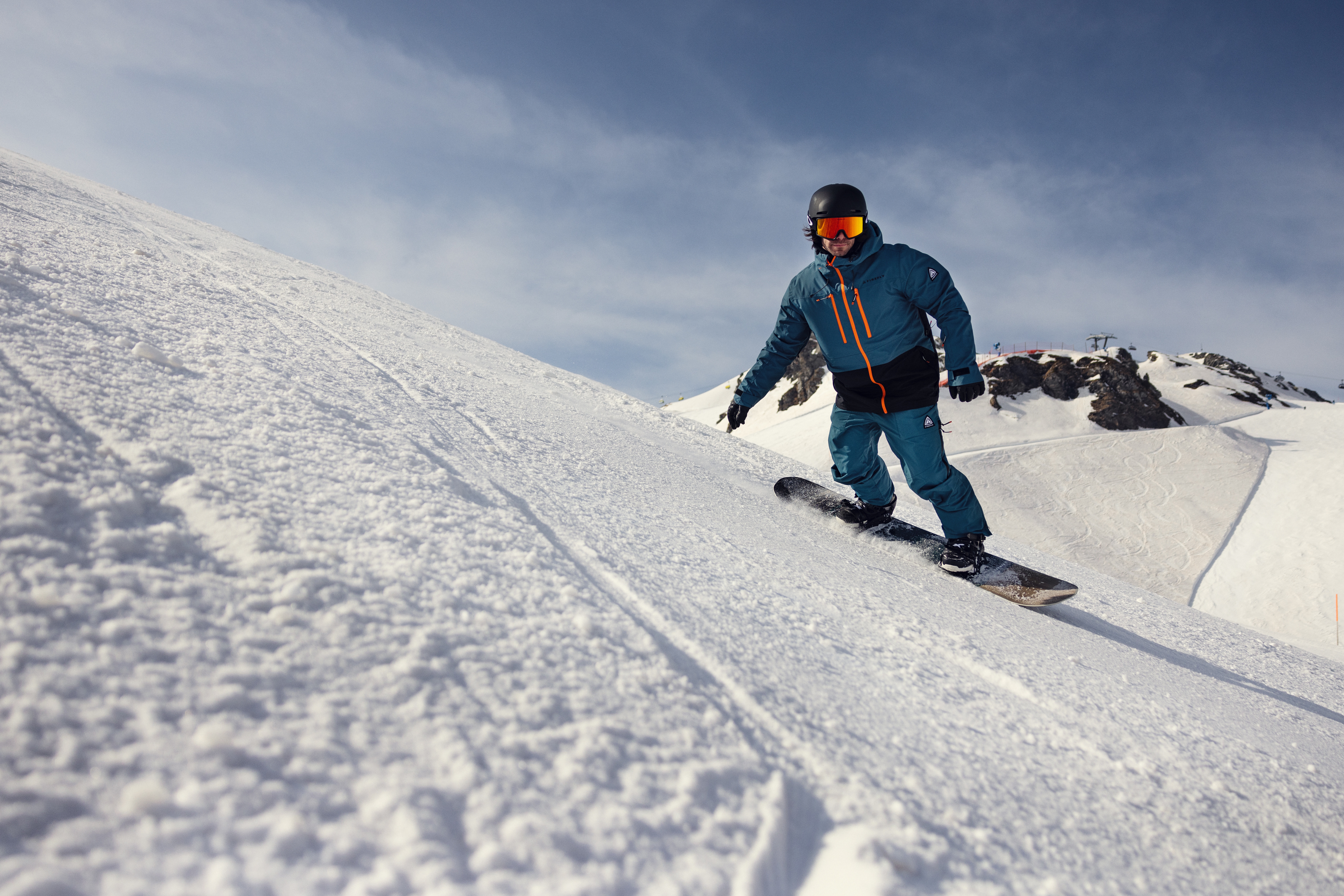 Ein Snowboarder in blauer Ausrüstung und schwarzem Helm gleitet unter strahlend blauem Himmel einen verschneiten Hang hinunter und vermittelt dabei das Gefühl von Aufregung und Freiheit in den Bergen.