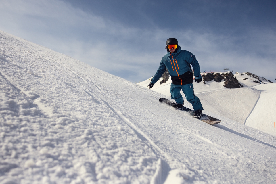 A snowboarder in blue gear and a black helmet glides down a snowy slope under a clear blue sky, conveying excitement and freedom in the mountains.