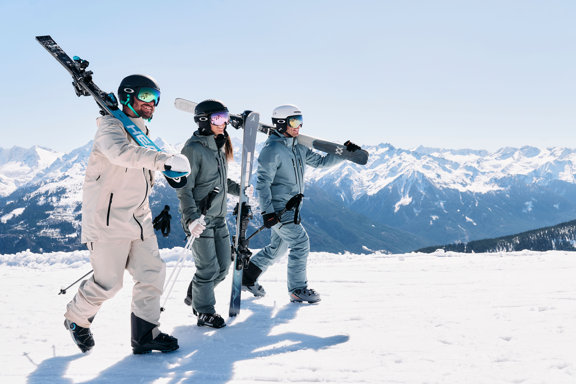 Three people in ski gear walk on a snowy mountain, carrying skis. Snow-capped peaks and a clear blue sky create a vibrant, adventurous atmosphere.