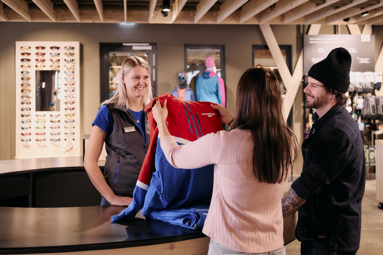 A woman holds a red and blue jacket at a store counter, smiling at the cashier. A man in a beanie stands beside her. The shop has a warm, inviting atmosphere.