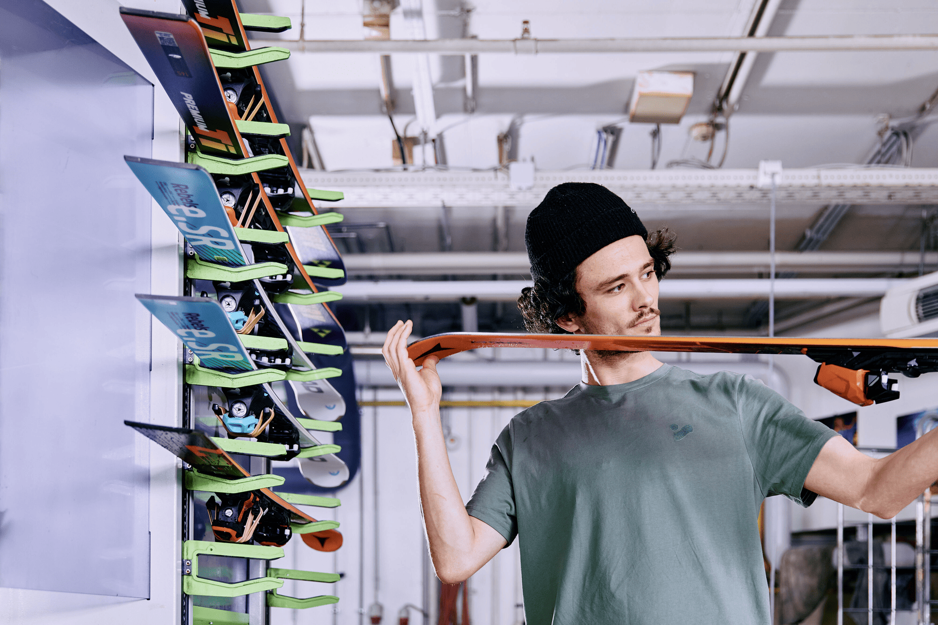 A young man in a beanie inspects a ski, holding it up horizontally. Behind him, a rack displays several colorful skis. The setting is industrial.