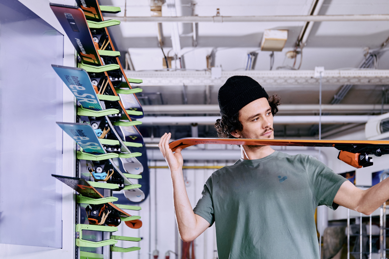 Ein junger Mann mit einer Mütze inspiziert einen Ski, indem er ihn horizontal hochhält. Hinter ihm sind auf einem Gestell mehrere bunte Skier ausgestellt. Die Umgebung ist industriell geprägt.