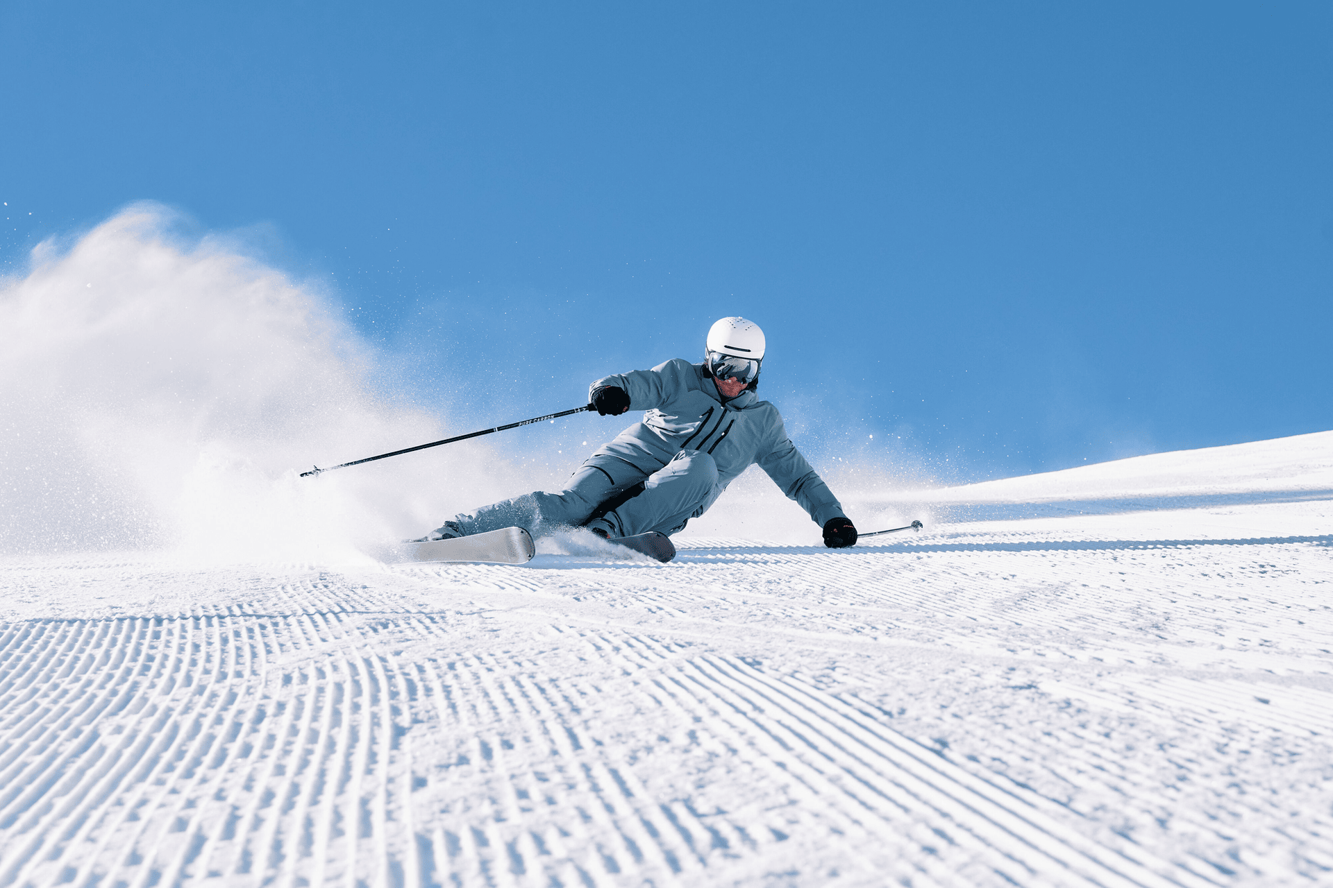 A skier in a blue suit and helmet carves smoothly down a sunlit slope, leaving a trail of snow with a clear blue sky in the background.