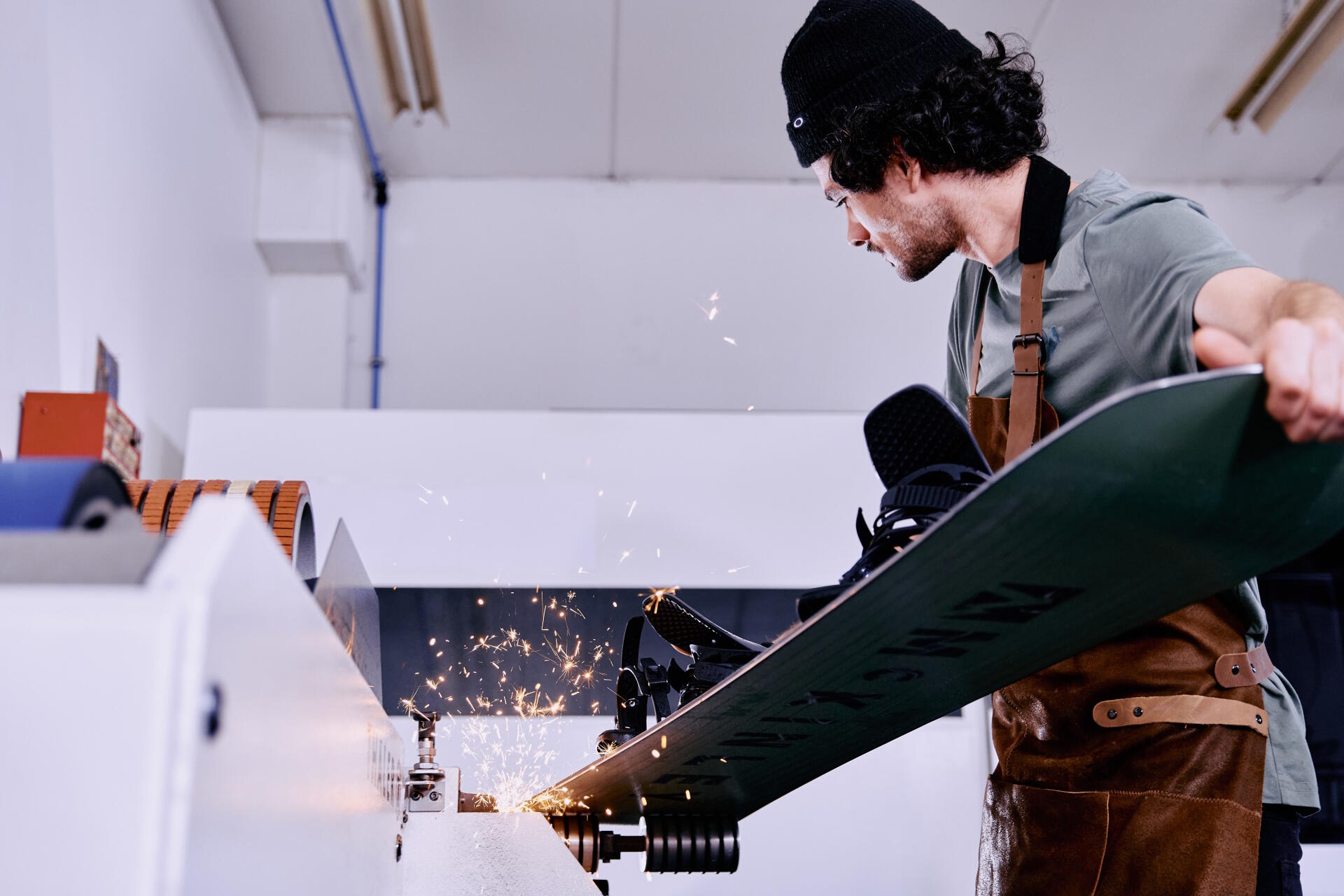 Ein Mann arbeitet in einer Skiwerkstatt und verwendet eine Maschine, um Skier zu tunen. In der Nähe stehen verschiedene Skier und ein Snowboard aufrecht, was Präzision und Konzentration suggeriert.
