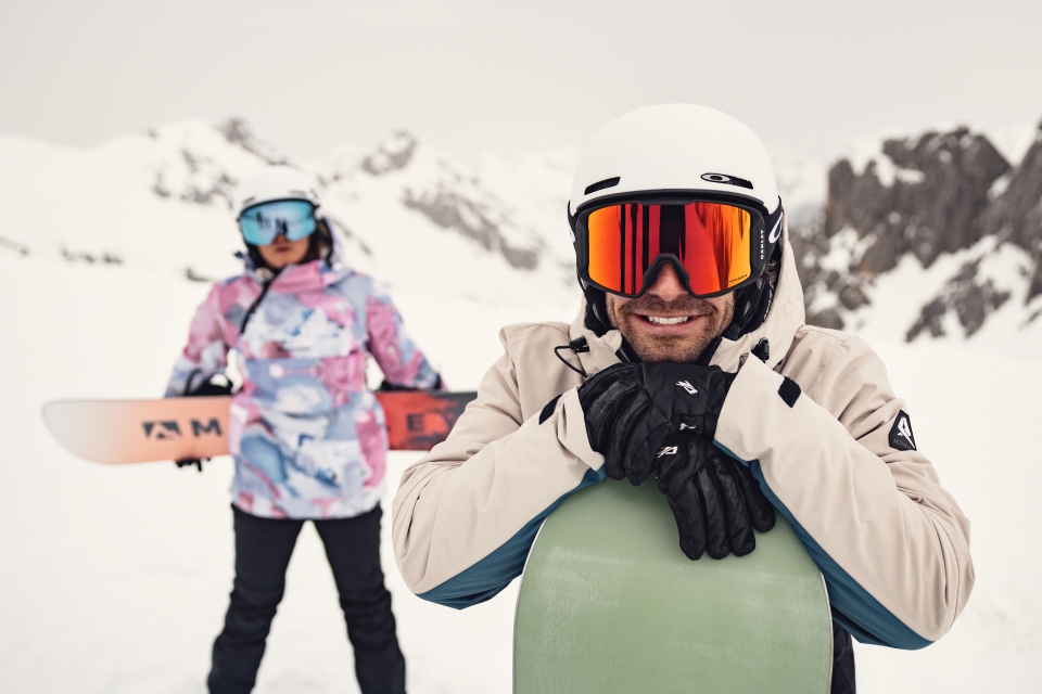 A smiling person leans on a snowboard in the foreground, wearing a white helmet and reflective goggles. Another person stands behind with a snowboard, wearing colorful gear. Snowy mountains form the backdrop.